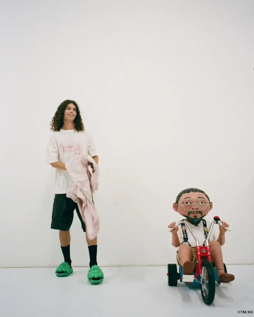 Wide studio image showing a model in an oversized white graphic T-shirt, black shorts, and bright green slippers standing beside a whimsical character sculpture riding a tricycle. The minimalist setting enhances the playful atmosphere