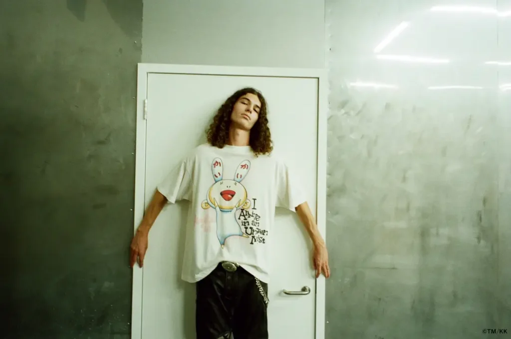 Model poses against a white door inside an industrial interior while wearing an oversized white graphic T-shirt with a cartoon rabbit illustration. Metallic walls and moody lighting add an experimental fashion-editorial aesthetic