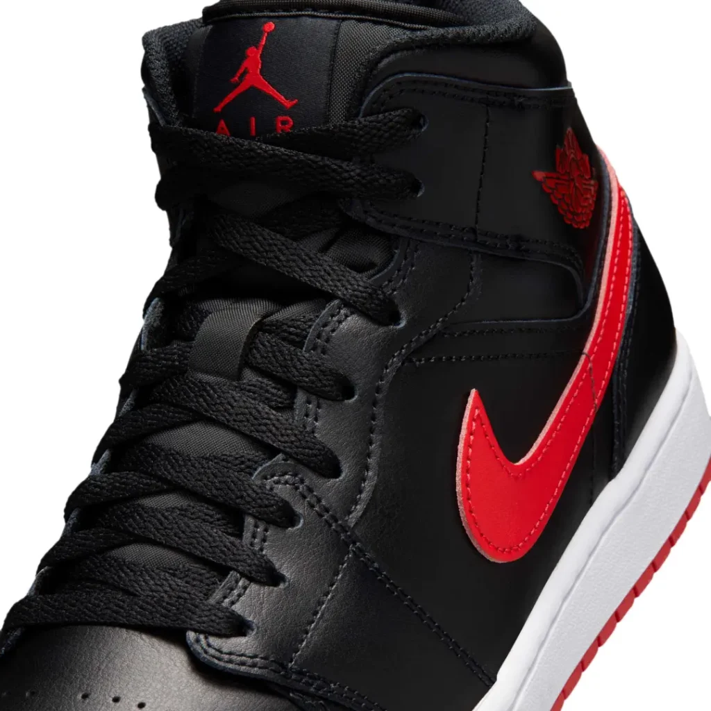 Close-up detail of the Air Jordan 1 Mid’s black leather upper, focusing on the bright red Swoosh, black woven laces, red Jumpman tongue branding, and embossed Wings logo near the ankle collar