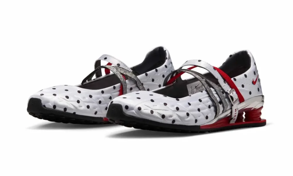 Pair of Nike Shox Mary Jane sneakers in white satin with black polka dots, photographed against a light gray studio background. The design features metallic silver crossover straps, red elastic detailing, chrome heel accents, and signature red Shox cushioning column