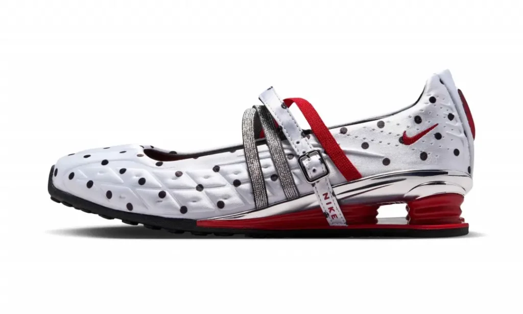 Side profile of a white Nike Shox Mary Jane shoe showcasing black polka-dot graphics, a red Swoosh, silver crisscross straps, adjustable buckle closure, and glossy chrome heel components above red Shox cushioning