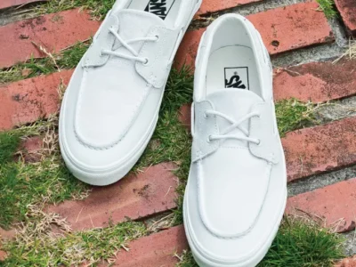 A pair of monochromatic white Vans boat shoes created exclusively for Urban Research DOORS is photographed from above on a weathered brick pathway lined with patches of green grass. The minimalist design combines suede-textured uppers, tonal laces, and clean white rubber soles, emphasizing a refined yet casual aesthetic. Branding appears on the insoles, while promotional text at the bottom highlights the collision
