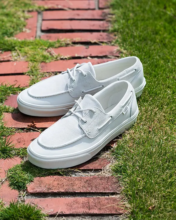 The Vans boat shoes are shown in a side-profile lifestyle shot outdoors on a sunlit brick-and-grass path. Soft suede overlays, moc-toe stitching, and wraparound lacing details define the silhouette, while the all-white color scheme gives the classic deck-shoe-inspired design a modern, understated look