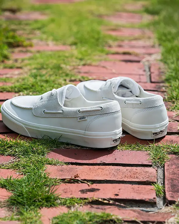 A rear three-quarter view showcases the pair resting on uneven red bricks surrounded by grass. Visible details include the heel construction, tonal stitching, side Vans tag, and signature “Off The Wall” heel badge. The bright outdoor lighting emphasizes the shoe’s clean materials and monochrome finish