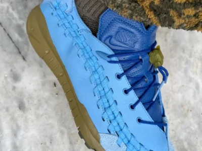 Close-up side profile of the Nike x SOULGOODS Homescape sneaker. The shoe combines a light blue leather upper with woven detailing along the sidewall, a darker blue tongue and laces, and a suede toe cap. An olive-green sole and translucent lace lock add contrast, while patterned fleece pants and marble flooring create a lifestyle-focused set