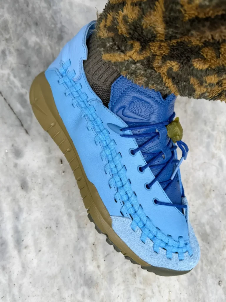 Close-up side profile of the Nike x SOULGOODS Homescape sneaker. The shoe combines a light blue leather upper with woven detailing along the sidewall, a darker blue tongue and laces, and a suede toe cap. An olive-green sole and translucent lace lock add contrast, while patterned fleece pants and marble flooring create a lifestyle-focused set