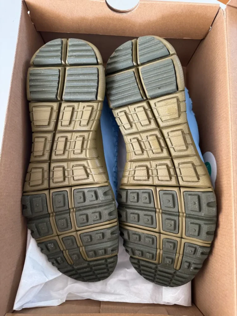Bottom view of both sneakers inside the box, revealing the segmented olive and gray rubber outsole. The tread pattern combines flexibility and traction, while speckled detailing reinforces the shoe’s outdoor-inspired, lifestyle-oriented design lang