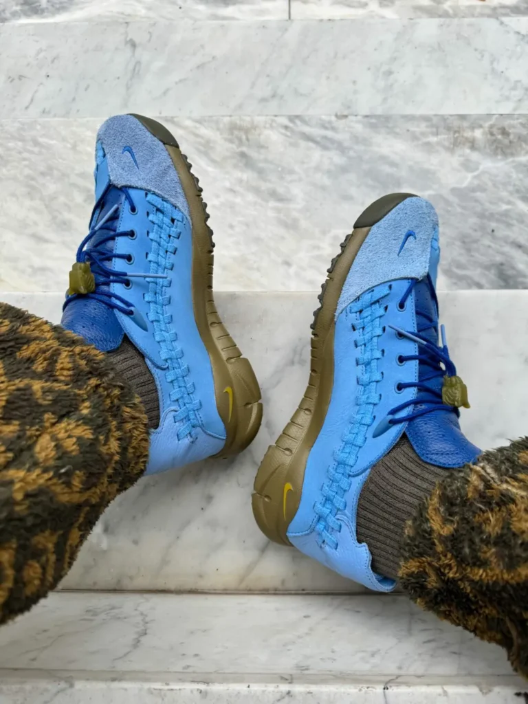 Top-down view of both shoe resting on marble stairs. The image highlights the symmetrical woven construction, dual-tone blue upper, olive midsoles, and small Swoosh branding on the toe boxes. The fleece pants frame the footwear from above