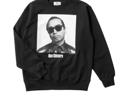Black crewneck sweatshirt featuring a large black-and-white portrait of Ken Shimura wearing dark sunglasses. His name is printed beneath the image in bold lettering, creating a minimalist memorial-inspired graphic on a heavyweight cotton sweatshirt with ribbed cuffs and hem