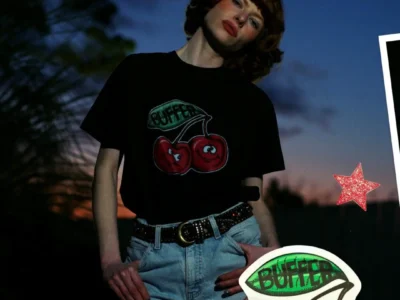 Editorial-style campaign image featuring a model in a black graphic T-shirt with a cartoon cherry logo, light-wash jeans, and a studded belt, photographed at dusk against a deep blue sky and framed by colorful sticker graphics and scrapbook-inspired design element