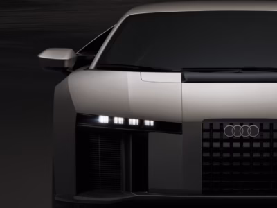 Matte silver Audi concept sports car with a squared-off front fascia, illuminated segmented LED headlights, deep vertical air intakes, black grille with Audi rings, and sharp geometric bodywork against a dark backdrop