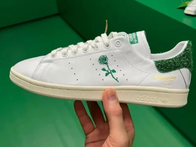 On-foot view of the collision's fully green shaggy-textured adidas Stan Smith. White laces, a green tongue label, cream sole, and white socks paired with white trousers highlight the grass-like upper and premium material