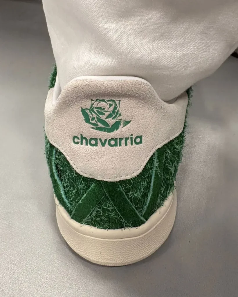Back angle of the white leather adidas Stan Smith featuring quilted heel stitching and a shaggy green heel overlay. A hand holds the shoe against a bright green background, emphasizing the textured materials and cream rubber sole
