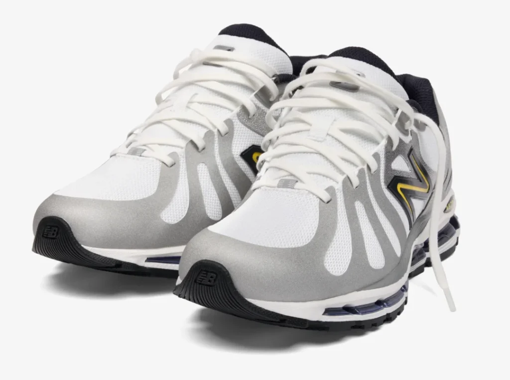 Front three-quarter view of the silver JJJJound x New Balance 1890 featuring metallic grey overlays, white mesh panels, yellow logo accents, and visible cushioning technology