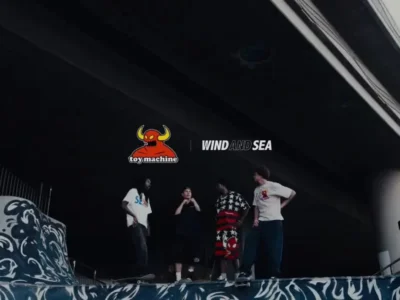 Group of skateboarders gather beneath an urban overpass at a skate spot decorated with bold wave-inspired graphics. The Toy Machine and WIND AND SEA collaboration logos appear above the group as several people wear graphic T-shirts and streetwear while standing with skateboard