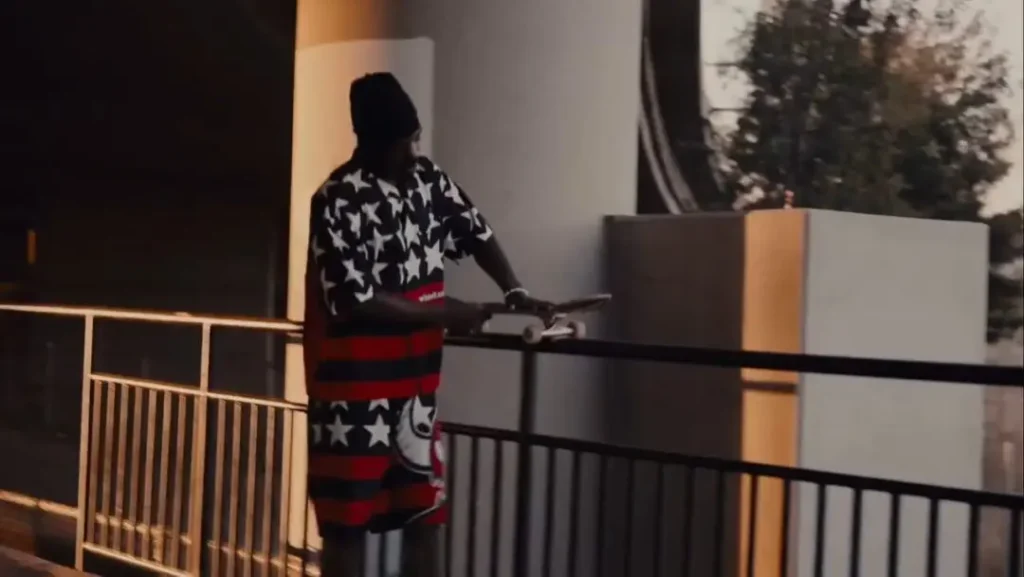 A skateboarder in a black beanie wears a matching oversized shirt and shorts set decorated with white stars, red stripes, and Toy Machine graphics while balancing beside a handrail. Warm evening light and an urban skate environment highlight the bold collide apparel