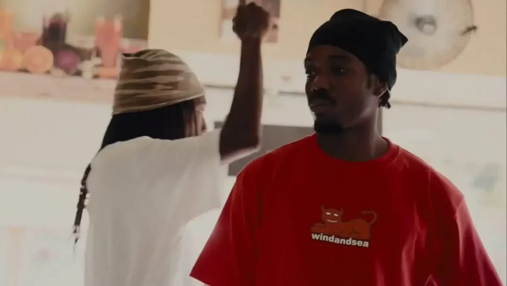 Two skateboarders stand inside a skate shop or indoor gathering space. The person in the foreground wears a bright red WIND AND SEA x Toy Machine graphic T-shirt featuring the iconic devil mascot, while another skater in a white shirt gestures in the softly lit background