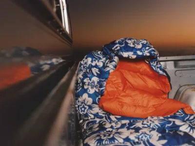 Blue floral-print sleeping bag with bright orange interior lining positioned in the back of a pickup truck at sunset, showcasing the Stüssy x Mountain Hardwear collaboration’s outdoor-focused design and bold color contrast