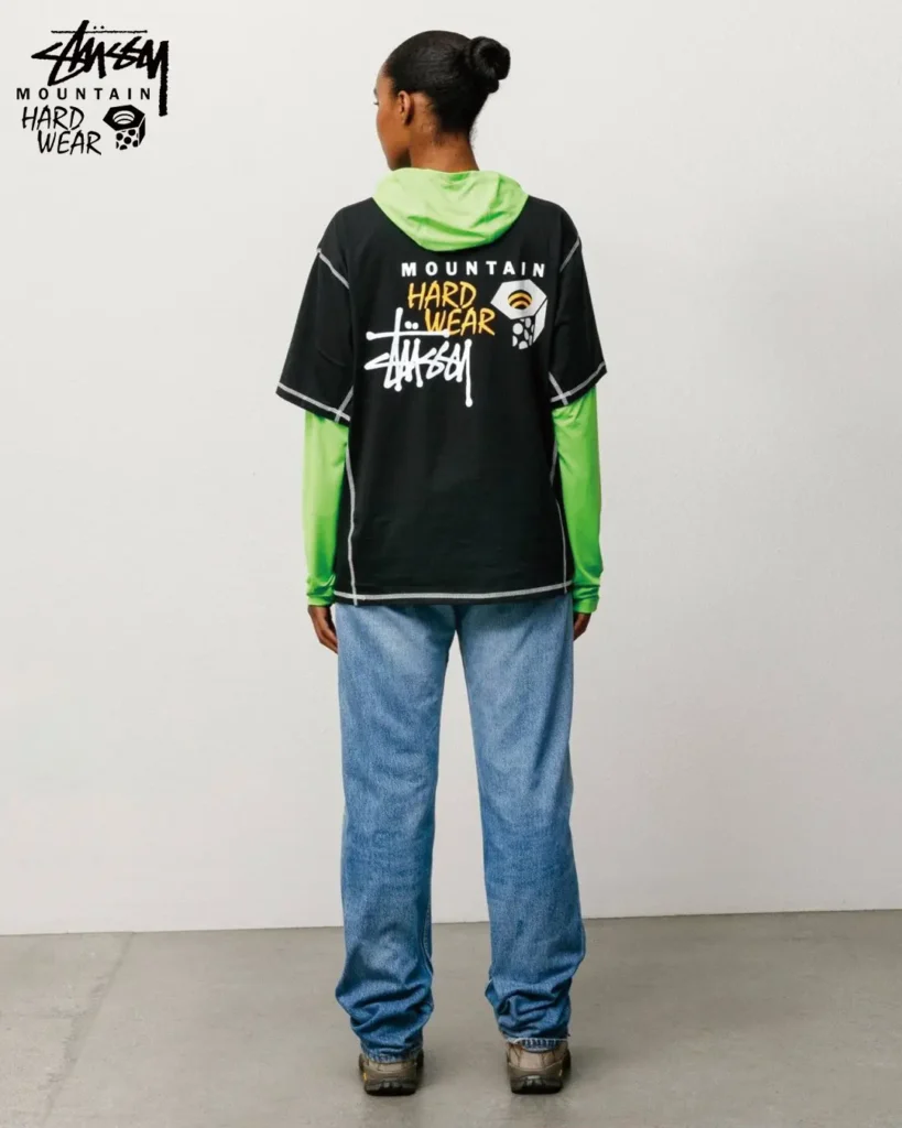Rear view of a black Stüssy x Mountain Hardwear performance T-shirt layered over a bright green hooded base layer with large collaborative artwork across the back