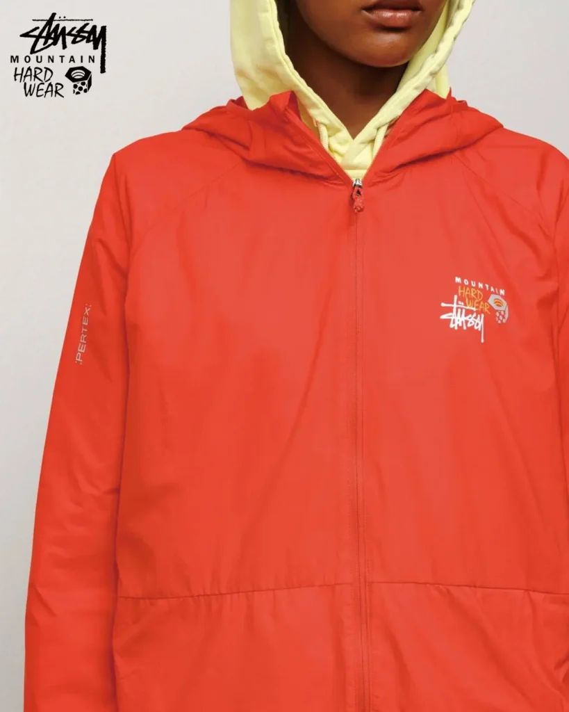 Close-up of a bright orange Pertex shell jacket from the Stüssy x Mountain Hardwear collection, featuring lightweight weather-resistant construction and co-branded chest graphic