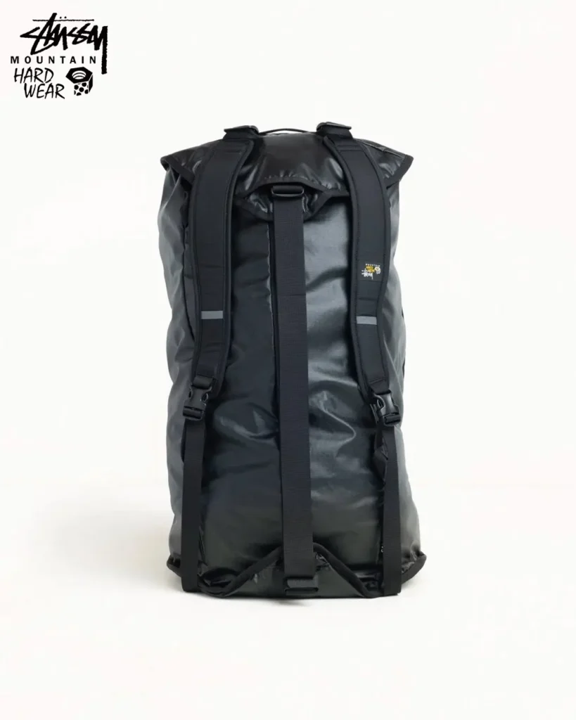 Rear view of a large black waterproof roll-top backpack from the Stüssy x Mountain Hardwear collection, featuring padded shoulder straps, reflective details, and expedition-ready storage capacity