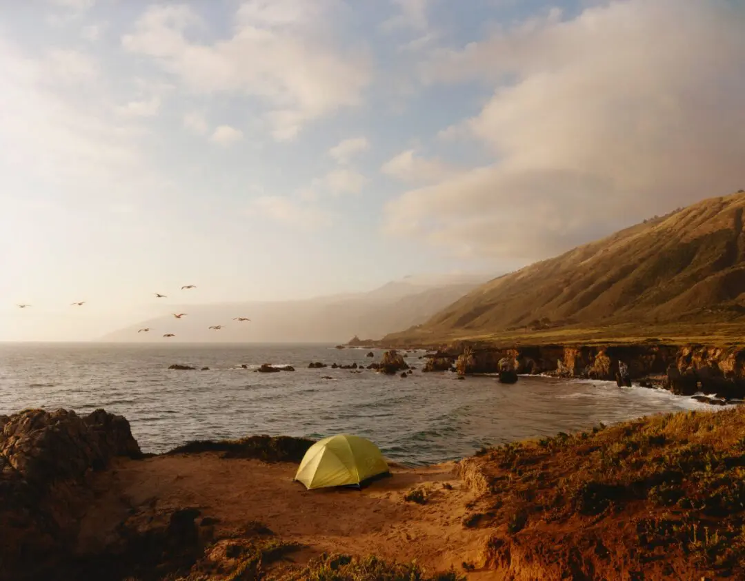 Coastal campsite overlooking rugged cliffs and ocean waves at golden hour, featuring a small tent on a scenic bluff that captures the adventurous spirit of the Stüssy x Mountain Hardwear collection