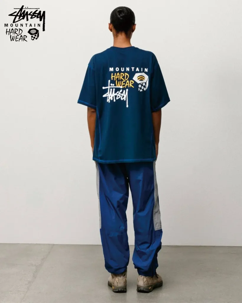Rear view of a model wearing a navy Stüssy x Mountain Hardwear graphic T-shirt paired with technical outdoor pants and hiking footwea