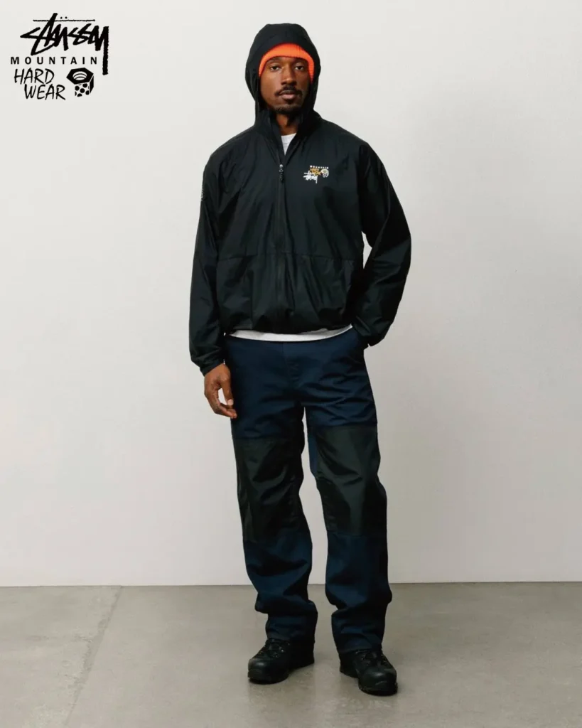 Model dressed in a black Stüssy x Mountain Hardwear hooded shell jacket and technical outdoor trousers, emphasizing lightweight weather protection and utility