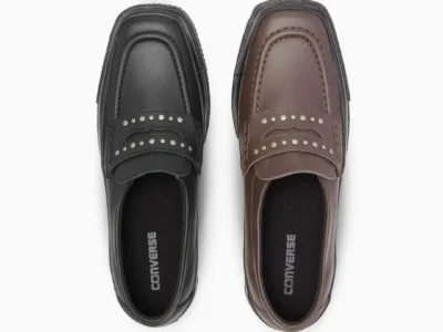 Black and brown Converse penny loafers displayed side by side, featuring square moc-toe construction, silver metal studs across the penny strap, smooth leather uppers, and black rubber Converse-inspired shoe