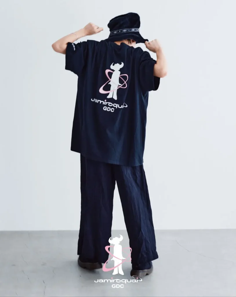 Product image of the black Jamiroquai x GDC T-shirt displayed against a colorful retro-futuristic radial background. The back graphic showcases the iconic buffalo-man silhouette with pink orbital accents and collide brand