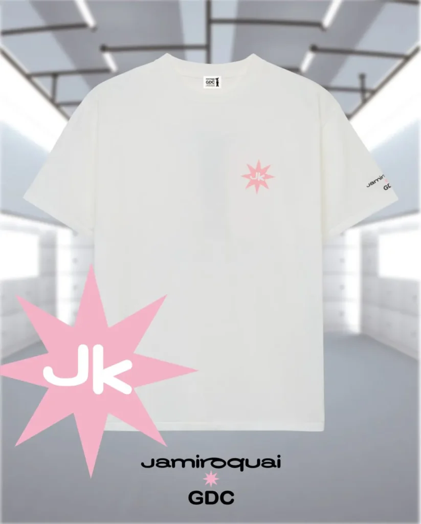 The oversized tee features a minimalist design with a small pink starburst graphic containing “JK” on the left chest, subtle co-branded sleeve detailing, and clean white cotton construction. A larger pink star motif and Jamiroquai x GDC branding appear below the garment, emphasizing the collection’s retro-futuristic music and streetwear influence