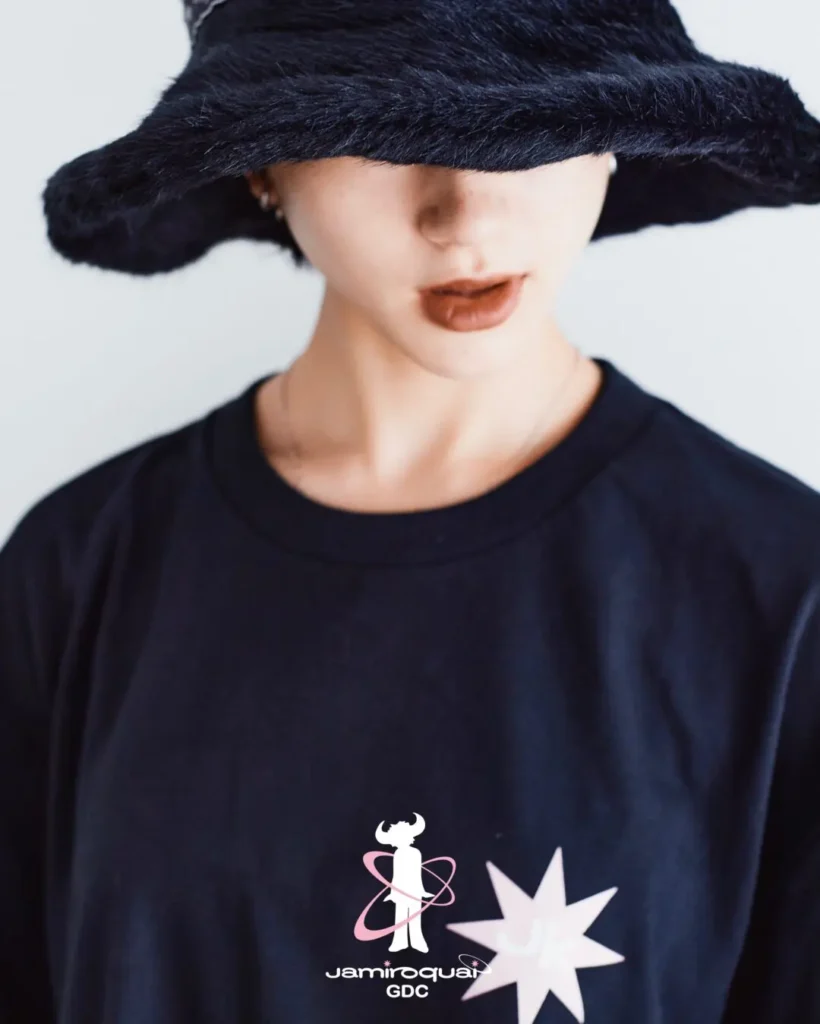 Close-up portrait of a model wearing a black Jamiroquai x GDC T-shirt and oversized fuzzy bucket hat. The shirt’s chest graphic combines the iconic buffalo-man logo with a soft pink orbital motif and starburst accent