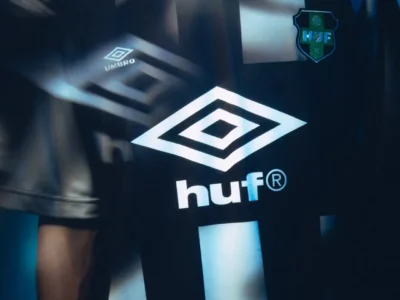 Close-up of a black HUF FC football jersey by Umbro featuring bold white vertical graphics, oversized Umbro diamond branding, HUF sponsor logo, and a club crest, photographed with motion blur and blue lighting for a futuristic sportswear campaign aesthetic