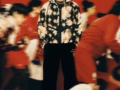 Model wearing a black Nike x PeaceMinusOne daisy-print jacket and matching athletic apparel, posed against a motion-blurred red studio background