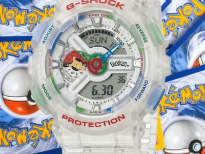 A close-up product shot of the Pokémon x G-SHOCK GA-110 collaboration watch featuring a transparent resin case and strap. The oversized analog-digital dial showcases colorful Pokémon-inspired details, including blue and green hands, multicolored accents, and a Pokémon logo at the center. Red “G-SHOCK” and “PROTECTION” branding stand out on the bezel, while translucent side buttons add a futuristic touch. The watch is displayed against a backdrop of scattered Pokémon trading cards and Poké Ball graphics, emphasizing the crossover’s nostalgic gaming appeal and collectible status