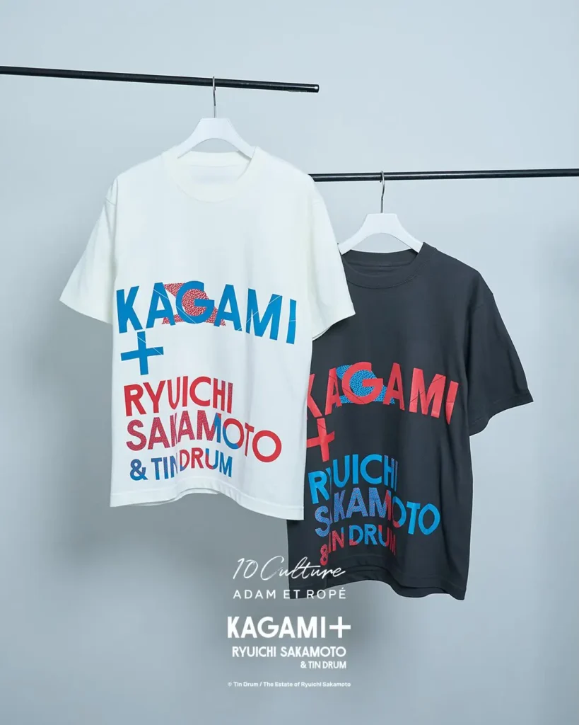 White and charcoal KAGAMI+ T-shirts feature bold red and blue typography spelling out Ryuichi Sakamoto and Tin Drum across oversized graphic layouts. The vibrant lettering contrasts with the minimalist garment color
