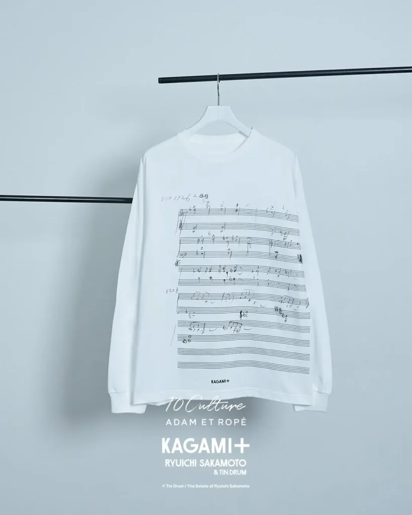White long-sleeve KAGAMI+ shirt featuring a large print of handwritten musical notation across the front. The minimalist design references Ryuichi Sakamoto's original composition sketches and creative process