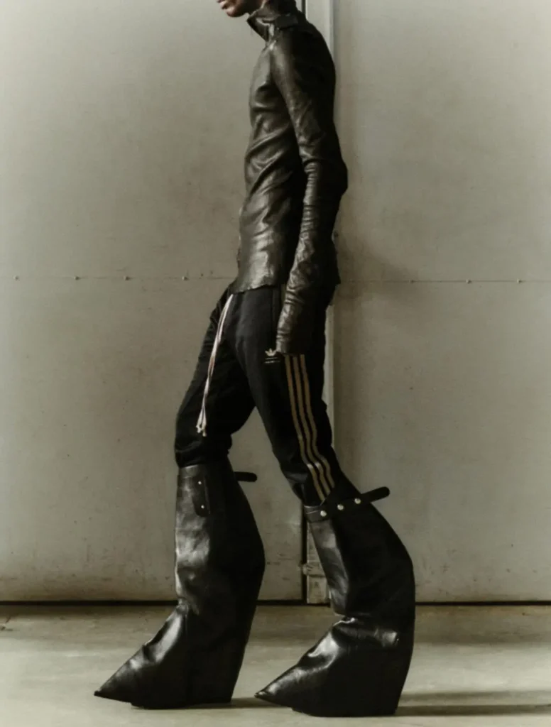 Side profile of a runway model wearing a fitted black leather top, adidas track pants with Three Stripes, and towering sculptural leather boots with exaggerated pointed toes. The industrial setting underscores the avant-garde style