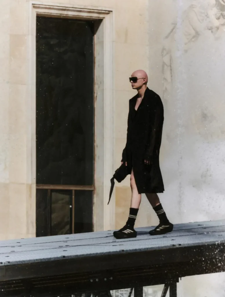 Portrait of a bald runway model wearing a black sleeveless draped top and adidas track pants with signature Three Stripes. Heavy black eye makeup and a dark backdrop create a stark, min editorial aesthetic