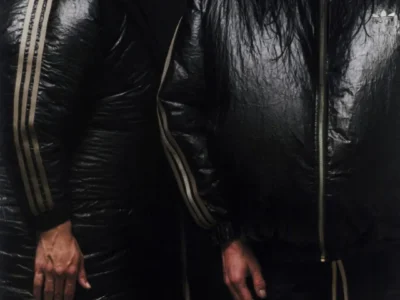 Studio portrait of two models wearing glossy black adidas Originals by Willy Chavarria outerwear. One model wears a long quilted coat with Three Stripes running down the sleeves, while the other wears a cropped insulated jacket with a high collar and matching shorts, emphasizing the collection's bold proportions and monochrome finish