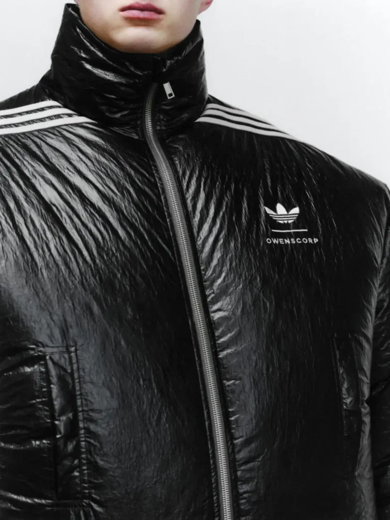 Close-up portrait of a model wearing an oversized glossy black insulated jacket from the adidas Originals x Willy Chavarria collection. The jacket features an exaggerated high collar, broad shoulders, Three Stripes across the upper chest and shoulders, and adidas Originals branding on the front