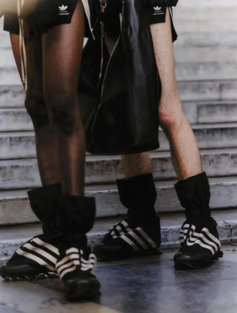 Side profile of a runway model wearing a fitted black leather top, adidas track pants with Three Stripes, and towering sculptural leather boots with exaggerated pointed toes. The industrial setting underscores the avant-garde style