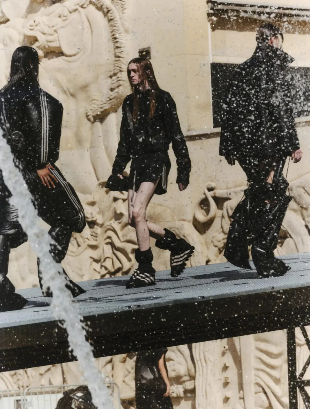 Models walk across an elevated outdoor runway in front of a fountain wearing black adidas Originals x Willy Chavarria looks. Water droplets fill the foreground while oversized footwear, layered silhouettes, and dramatic tailoring create a cinematic fashion show