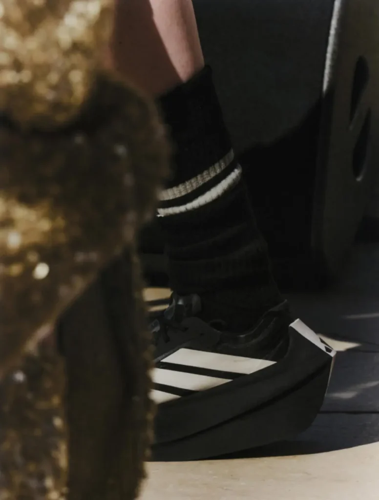 Close-up of a black adidas sneaker with oversized platform tooling and white Three Stripes paired with ribbed black socks. Warm sunlight highlights the geometric sole profile and minimalist style