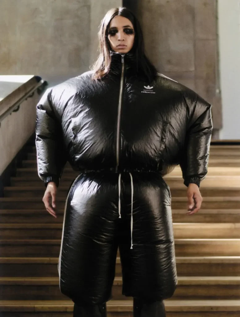 Front-facing runway portrait of a model wearing an exaggerated glossy black cropped puffer jacket and matching oversized quilted shorts. Dramatic black eye makeup and an industrial staircase backdrop enhance the futuristic sil
