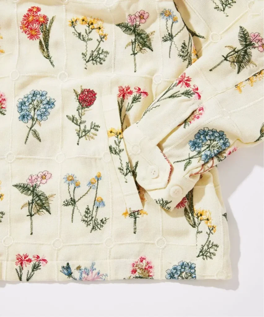 Macro imagery emphasizes the intricate embroidery techniques used throughout the garment. Colorful botanical illustrations emerge from the textured cream backdrop, showcasing the depth, texture, and artisanal character of the stitching