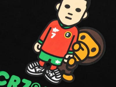 Close-up of the A BATHING APE x CR7 LIFE graphic featuring a cartoon-style Cristiano Ronaldo in a red and green football kit standing beside Baby Milo on a black Tee