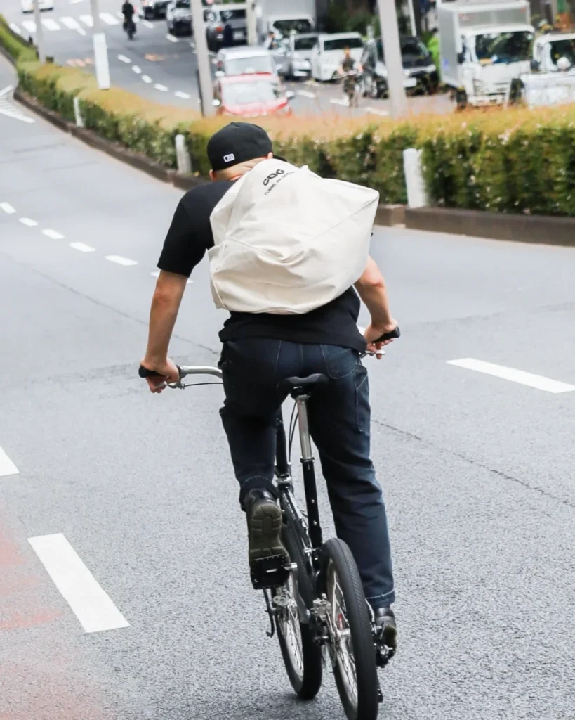 A person leans against a metal railing while carrying a black COMME des GARÇONS CDG tote bag over the shoulder, showcasing the bag’s relaxed drape and oversized design