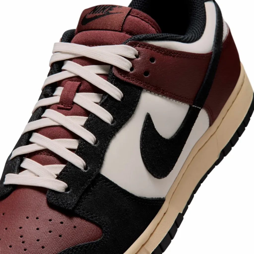 Close-up detail of the burgundy Nike Dunk Low highlighting cream flat laces, burgundy canvas tongue, black suede eyestays and Swoosh, sail side panel, and textured material finish