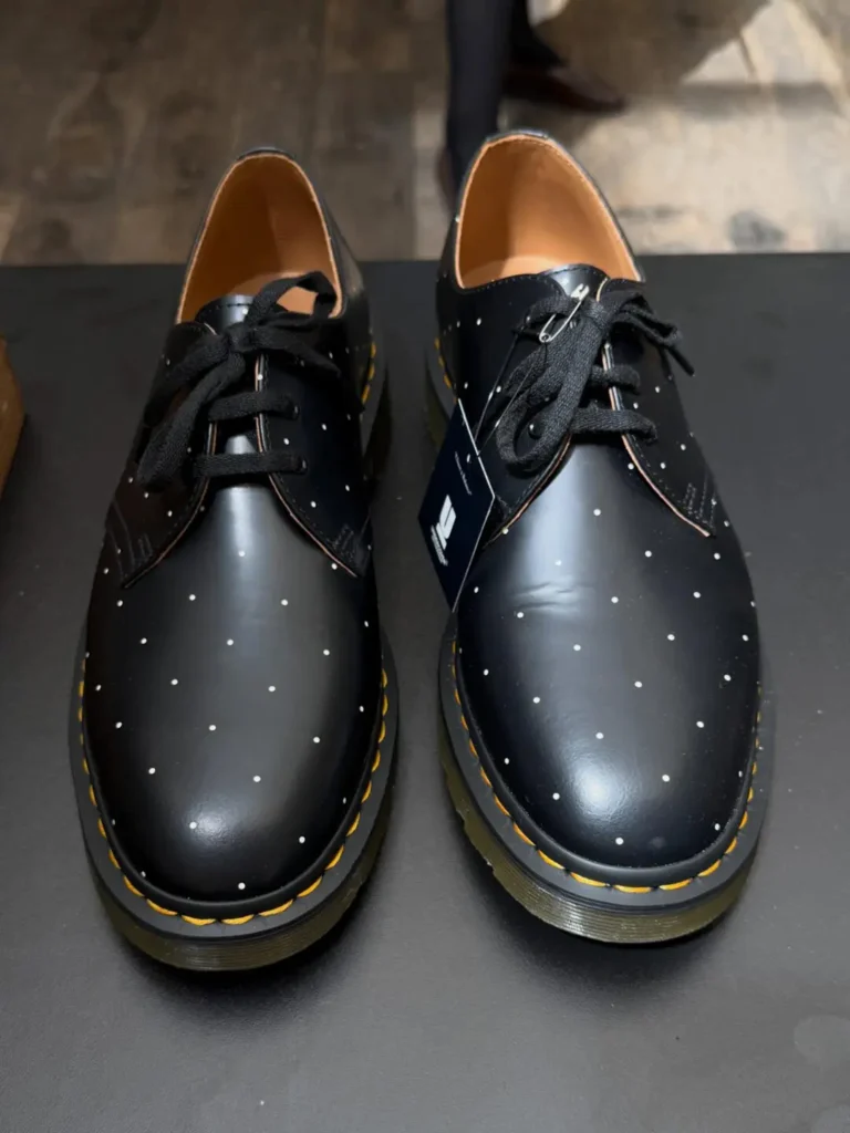Pair of black Dr. Martens 3-eye derby shoes displayed on a tabletop, finished with a subtle white polka-dot pattern, black laces, smooth leather uppers, signature yellow welt stitching, and translucent AirWair sole